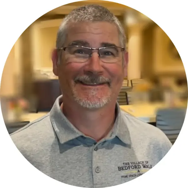 Close-up portrait of a smiling man with gray hair and glasses wearing a light gray polo shirt, blurred indoor background.