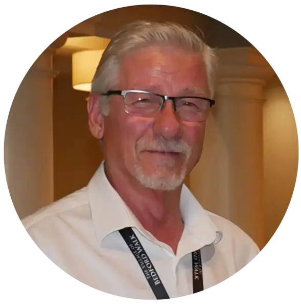 Portrait of an older man with gray hair and glasses, smiling, wearing a white shirt and a conference lanyard around his neck.
