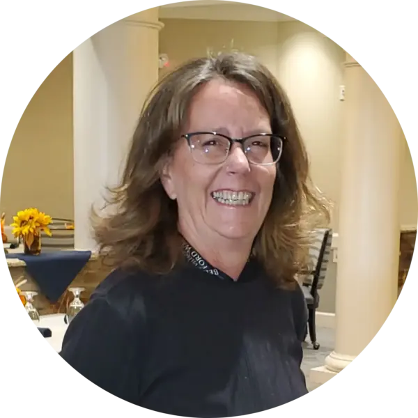 Smiling woman with glasses in a black top at an indoor event; flowers and tables in the background.